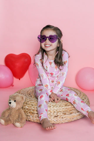 Child wearing pink pajamas with a teddy bear and heart pattern, sitting on a wicker stool with balloons in the background.
