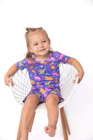 A young girl with her hair in a topknot sits on a white wire chair wearing a purple short-sleeve pajama set with a colorful cartoon animal print. The background is a clean white studio backdrop.