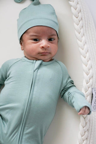 Baby in a light blue outfit and hat lying on a white surface with a textured white blanket.